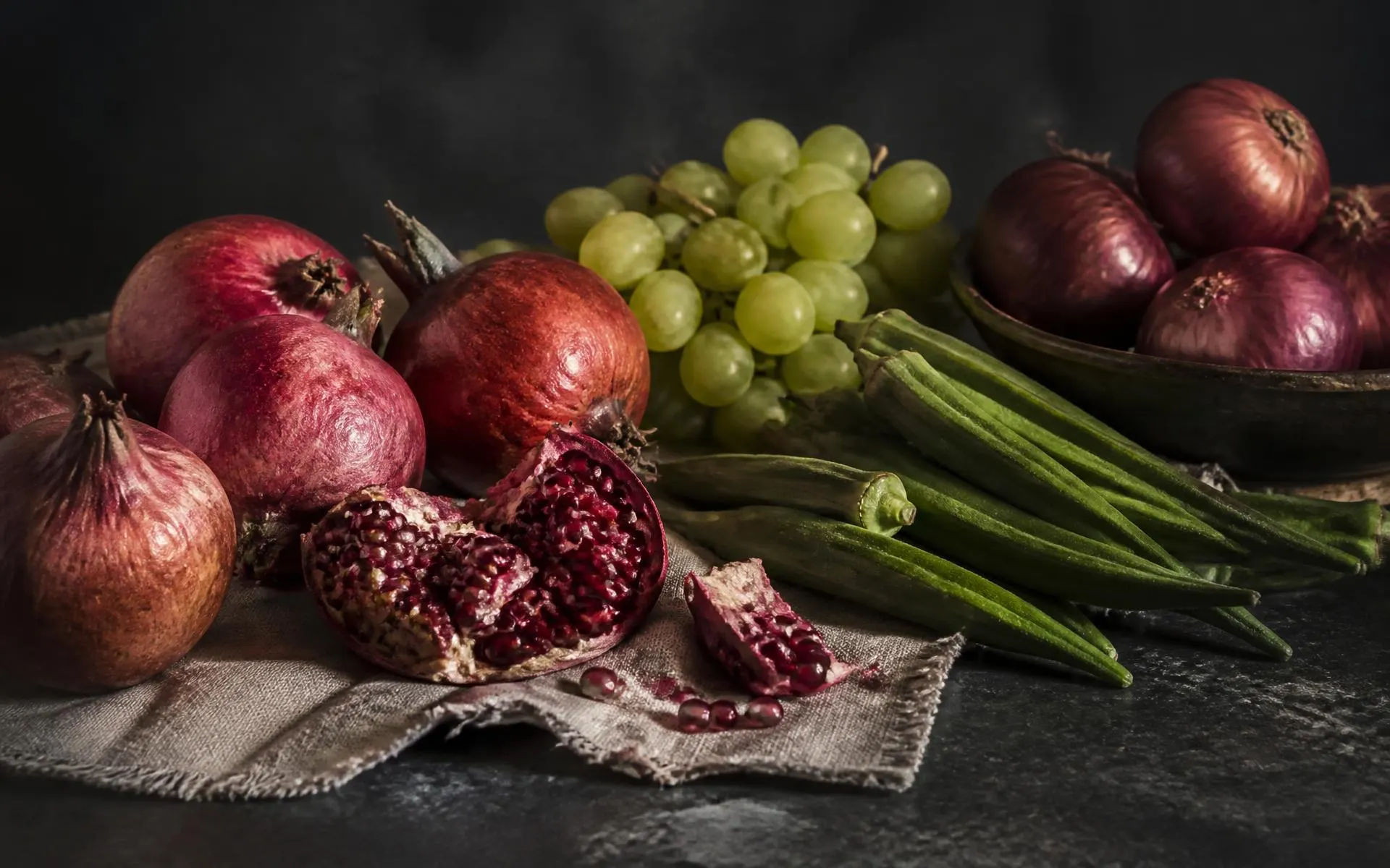 Pomegranates, grapes, okra and onions arranged on linen over dark stone