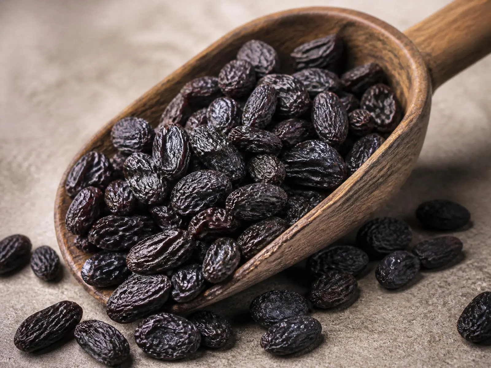 Dark black brown raisins in a wooden scoop on a neutral stone surface